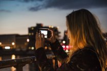 Young woman by waterfront, at dusk, photographing view with smartphone — Stock Photo