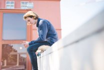 Giovane skateboarder urbano maschile seduto su skateboard lettura smartphone testo — Foto stock
