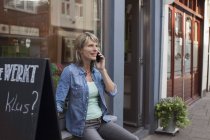 Mujer sentada en el alféizar de la ventana de la tienda haciendo llamada telefónica - foto de stock