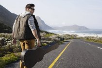 Vista trasera del joven caminando por la carretera costera llevando estuche de guitarra, Ciudad del Cabo, Cabo Occidental, Sudáfrica - foto de stock