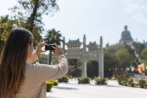 Joven turista tomando fotografías de teléfonos inteligentes de Tian Tan Buddha, Monasterio Po Lin, Isla Lantau, Hong Kong, China - foto de stock