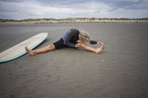 Senior woman sitting on sand, stretching, surfboard beside her — Stock Photo