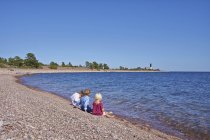 Drei Kinder sitzen am felsigen Strand bei blauem Wasser — Stockfoto
