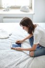 Молодая женщина сидит на кровати и читает цифровой планшет — стоковое фото
