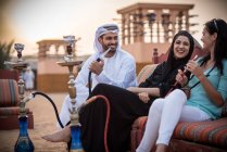 Casal local vestindo roupas tradicionais fumando shisha no sofá com turista feminino, Dubai, Emirados Árabes Unidos — Fotografia de Stock