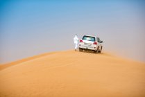 Homem do Oriente Médio vestindo roupas tradicionais com veículo off-road estacionado na duna do deserto, Dubai, Emirados Árabes Unidos — Fotografia de Stock
