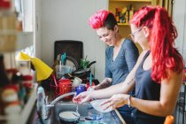 Дві молоді жінки з рожевим волоссям сміються під час миття посуду на кухні — стокове фото
