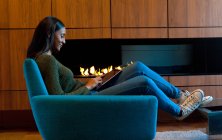 Woman in armchair in front of wooden cabinets and open fire, using digital tablet — Stock Photo
