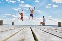 Young woman sitting on post on wooden pier, watching friends as they jump into sea — Stock Photo