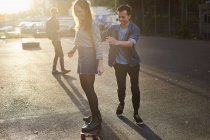 Jovem empurrando jovem skatista feminina na rua iluminada pelo sol — Fotografia de Stock