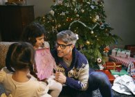 Padre y dos hijas abren regalos de Navidad en la sala de estar - foto de stock