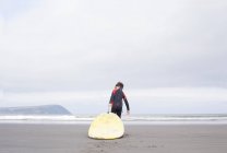 Vista trasera del niño tirando de la tabla de surf en la playa - foto de stock
