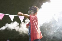 Niño con gafas y capa en posición de superhéroe frente a la nube de humo - foto de stock