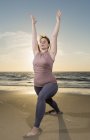 Mulher madura praticando ioga em uma praia ao pôr do sol, pose de guerreiro — Fotografia de Stock