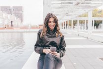 Молодая женщина сидит на улице, используя смартфон, улыбаясь — стоковое фото