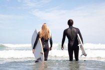 Pareja joven caminando hacia el mar sosteniendo tablas de surf, vista trasera - foto de stock