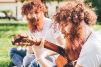 Joven macho hipster gemelos con barba roja sentado en el parque tocando la guitarra - foto de stock