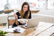 Jovem mulher no apartamento da cidade comendo café da manhã muesli enquanto lia laptop — Fotografia de Stock