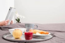 Breakfast tray on bed and person reading newspaper on background — Stock Photo