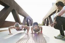 Two women doing push ups on urban footbridge with male personal trainer — Stock Photo