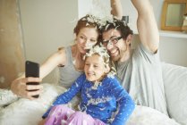 Casal adulto médio e filha coberta de penas de luta de travesseiro tomando selfie smartphone na cama — Fotografia de Stock