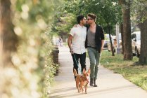 Jovem casal masculino beijando, andando cão na calçada suburbana — Fotografia de Stock