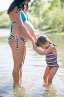 Madre embarazada e hija de pie en el lago, cogidas de la mano - foto de stock