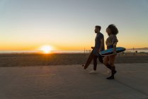 Casal andando ao longo do caminho pela praia, segurando skates — Fotografia de Stock