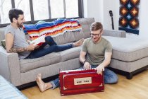 Мужская пара дома, молодой человек на диване смотрит, как его партнер играет на музыкальном инструменте — стоковое фото