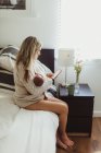 Доросла жінка сидить на ліжку і дивиться на смартфон, поки хитає новонародженою донечкою. — стокове фото