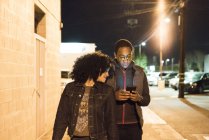 Couple walking in street at night looking at smartphone, Los Angeles, California, Usa. - foto de stock
