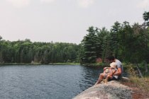 Joven y su hija sentados en la roca a la orilla de un lago - foto de stock