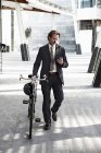 Mid adult businessman walking with bicycle and using mobile phone — Stock Photo