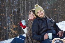Retrato de pareja sentada en la nieve con bebidas calientes - foto de stock