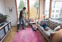 Man vacuuming carpet, girlfriend using laptop on sofa — Stock Photo