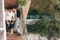 Joven pareja masculina abrazándose en el balcón del apartamento, leyendo el teléfono inteligente - foto de stock