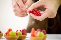 Male baker hands holding red currant over fruit tarts on table — Stock Photo