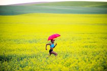 Молодая женщина бежит на поле рапса с красным зонтиком — стоковое фото