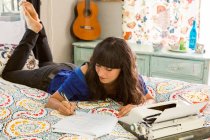 Молода жінка лежить на ліжку, використовуючи друкарську машинку, пишучи нотатки — стокове фото