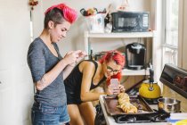 Duas jovens mulheres com cabelo rosa tirando fotografias de smartphones de baguete de pelúcia na cozinha — Fotografia de Stock