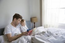 Padre e hijo pequeño, sentados en la cama, leyendo el libro juntos - foto de stock