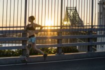 Jovem mulher tatuada correndo na ponte com o pôr do sol atrás — Fotografia de Stock