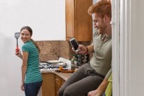 Молодий чоловік на кухні використовує смартфон, щоб сфотографувати молоду жінку, що тримає шпатель — стокове фото