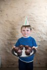Retrato de menino vestindo chapéu de festa segurando cupcakes — Fotografia de Stock