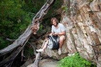 Retrato de un joven excursionista fotografiando sentado en la roca - foto de stock