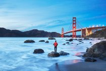 Vista panorámica del puente Golden Gate al atardecer con una persona parada en Marshall 's Beach en primer plano. San Francisco, California, Estados Unidos - foto de stock