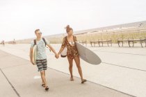 Молодая пара, держащаяся за руки и несущая доски для серфинга в Рокавей-Бич, штат Нью-Йорк, США — стоковое фото