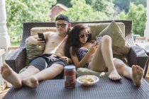 Підліток пара в купальних костюмах на ганку їсть закуски і дивиться на смартфони — стокове фото