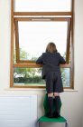 Visão traseira do menino da escola em pé em uma cadeira e inclinando-se para fora de uma janela de sala de aula — Fotografia de Stock