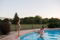 Молода дівчина стоїть на краю басейну, батько в басейні заохочує її стрибати — стокове фото
